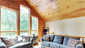 A cozy cabin-style room with two grey sofas, a wooden ceiling, and large windows offering a view of the forest. - Eagle on 7 by Taylor Made Deep Creek (McHenry)