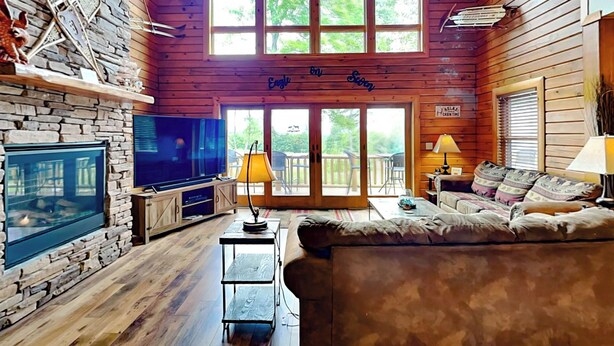 A cozy cabin interior with a stone fireplace, a large TV, a sofa, and a balcony with a view of trees. - Eagle on 7 by Taylor Made Deep Creek (McHenry)
