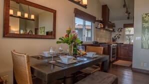 Comedor con una gran mesa de madera dispuesta para cuatro personas, rodeada de sillas de mimbre. Un espejo y lámparas colgantes están sobre la mesa.