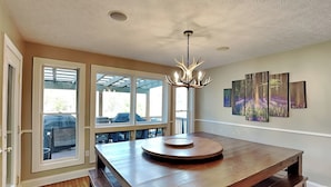 A spacious dining area with a large wooden table and benches, a chandelier, and a view through large windows to an outdoor patio.