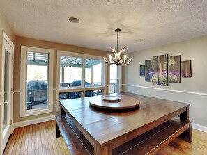 A spacious dining area with a large wooden table and benches, a chandelier, and a view through large windows to an outdoor patio. - Ever After by Taylor Made Deep Creek (Oakland)