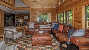 A log cabin interior with a large leather sofa, a patterned rug, and a dining area with wooden tables and chairs. - Family Sanity by Taylor Made Deep Creek (McHenry)