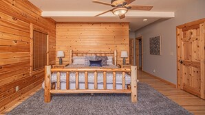 A wooden-paneled bedroom with a bed, two bedside lamps, a ceiling fan, and a door on the right. - Family Sanity by Taylor Made Deep Creek (McHenry)