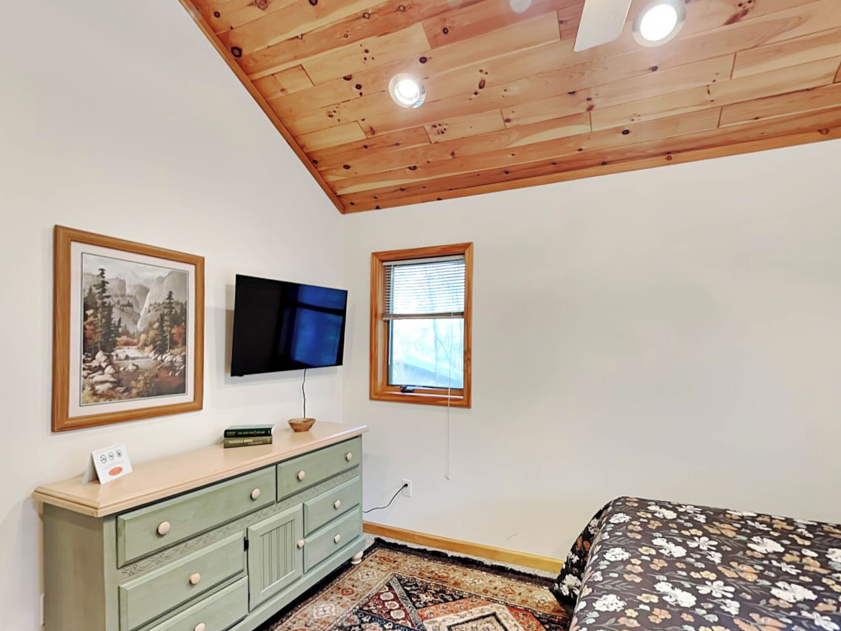 A bedroom with a wooden ceiling, a television mounted on the wall, a window with blinds, a bed with a floral bedspread, and a green dresser with drawers.