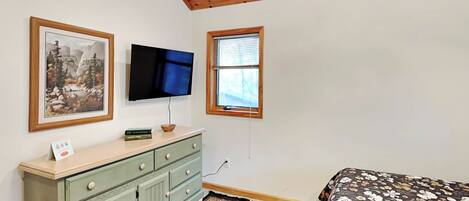A bedroom with a wooden ceiling, a television mounted on the wall, a window with blinds, a bed with a floral bedspread, and a green dresser with drawers.
