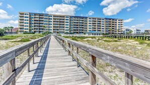 Un muelle de madera que lleva a un hotel de varios pisos con balcones, ubicado en una playa con poca vegetación.