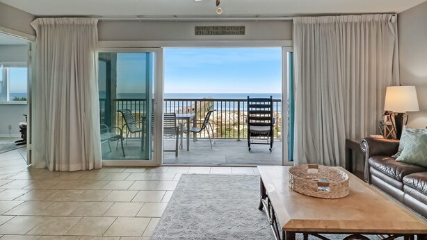 Una sala de estar con vista a la playa, un ventilador de techo, un letrero que dice "Life is Better at the Beach" y un comedor con sillas y una mesa.