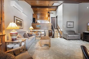 Amplia sala de estar con techo alto, vigas de madera, una escalera y un rincón de descanso con sofá y sillones.