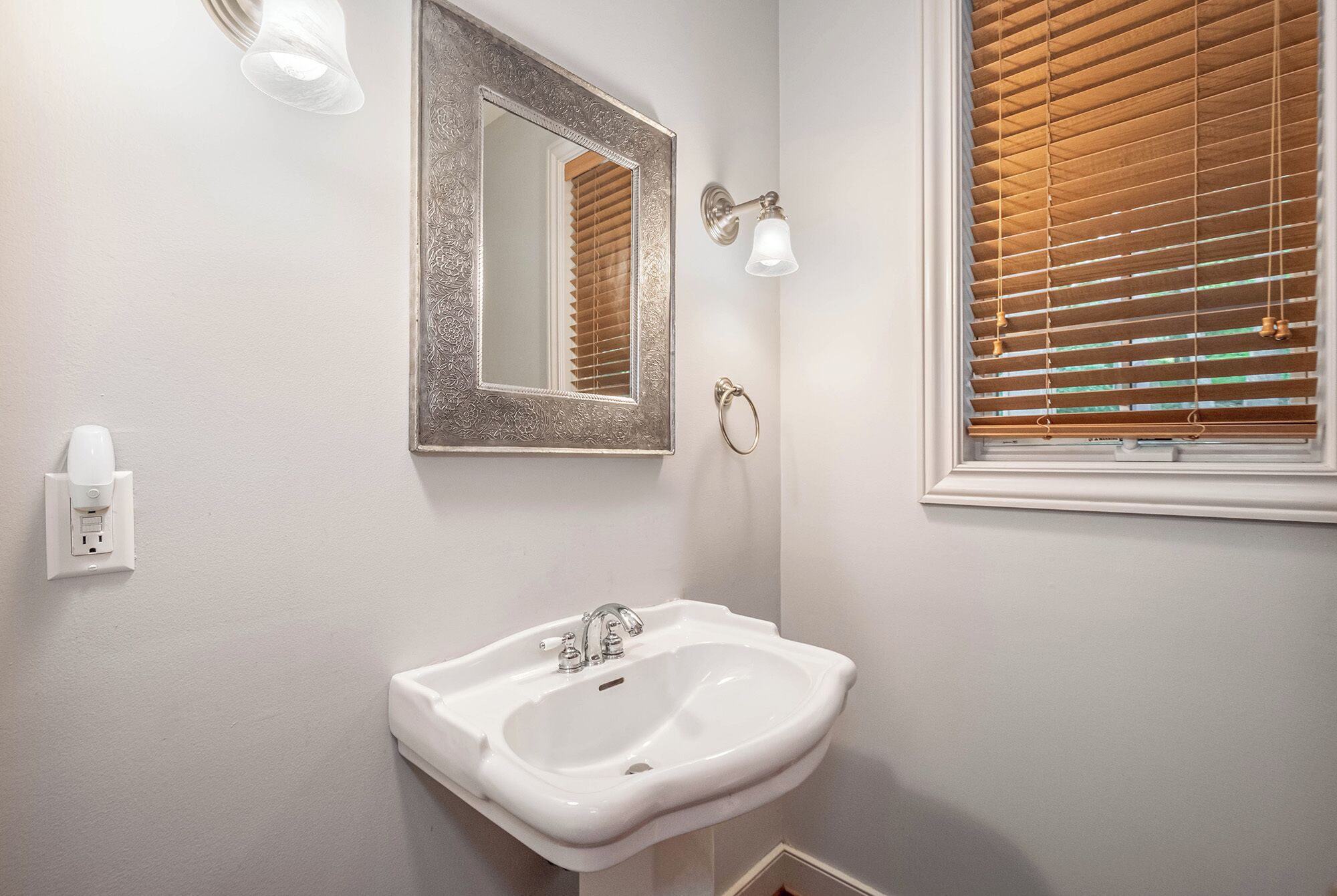 A bathroom with a white sink, a mirror with an ornate frame, a wall-mounted light, and a window with blinds.