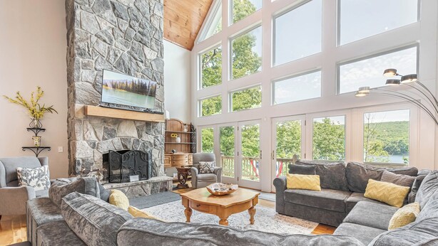 A spacious living room with a stone fireplace, large windows, and grey upholstered furniture. - Friday's Escape by Taylor Made Deep Creek (McHenry)