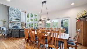 A spacious dining area with a large wooden table and chairs, a stone fireplace, and large windows offering views of the outdoors. - Friday's Escape by Taylor Made Deep Creek (McHenry)