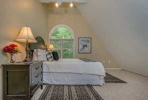 A bedroom with a bed, a dresser, a lamp, a window with a view of greenery, and a framed picture on the wall.