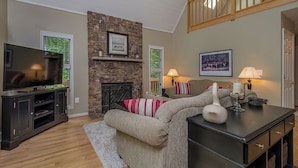 Una sala de estar con chimenea de piedra, un televisor sobre un mueble, un sofá con almohadones a rayas y una lámpara sobre una mesa.