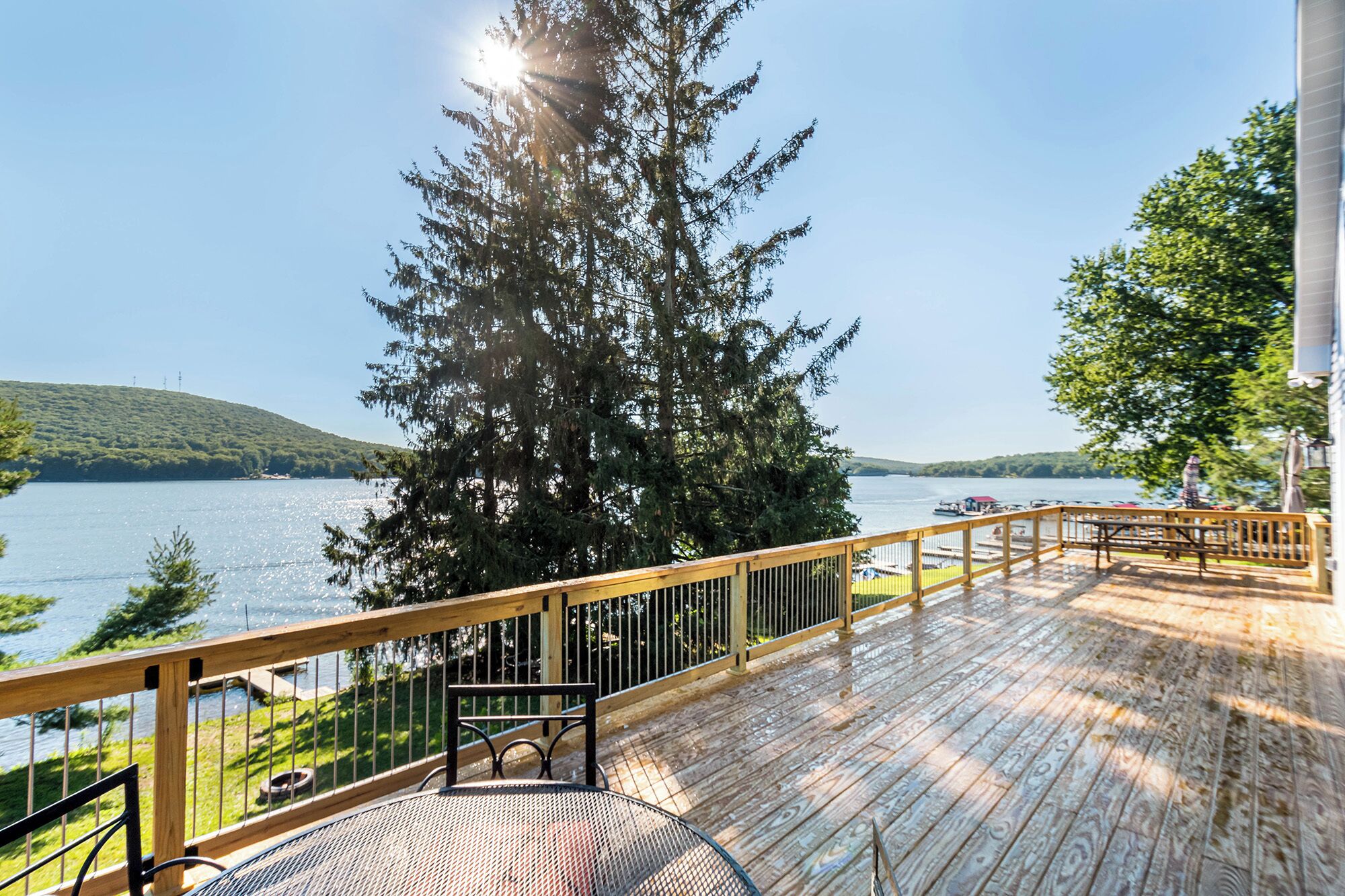 Terraza de madera con una mesa y sillas de metal, con vista a un lago con barcos y una colina boscosa al fondo.