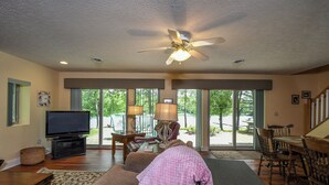A spacious living room with a ceiling fan, a television, a dining area with wooden chairs, and large windows offering a view of the outdoors. - Golden Pond by Taylor Made Deep Creek (Swanton)