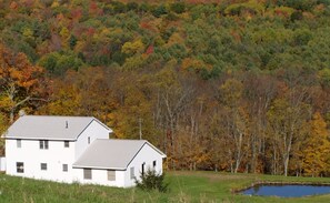 A two-story white house with a sloped roof, surrounded by a grassy area and a pond. - The Farmhouse, where peace and quiet awaits (Otego)