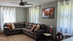 A living room with a sectional sofa, a chair, a table, a ceiling fan, and a painting on the wall.