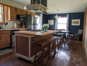 A kitchen with wooden cabinets, a stainless steel refrigerator, a gas stove, and a dining area with wooden chairs and a table. - The Farmhouse, where peace and quiet awaits (Otego)