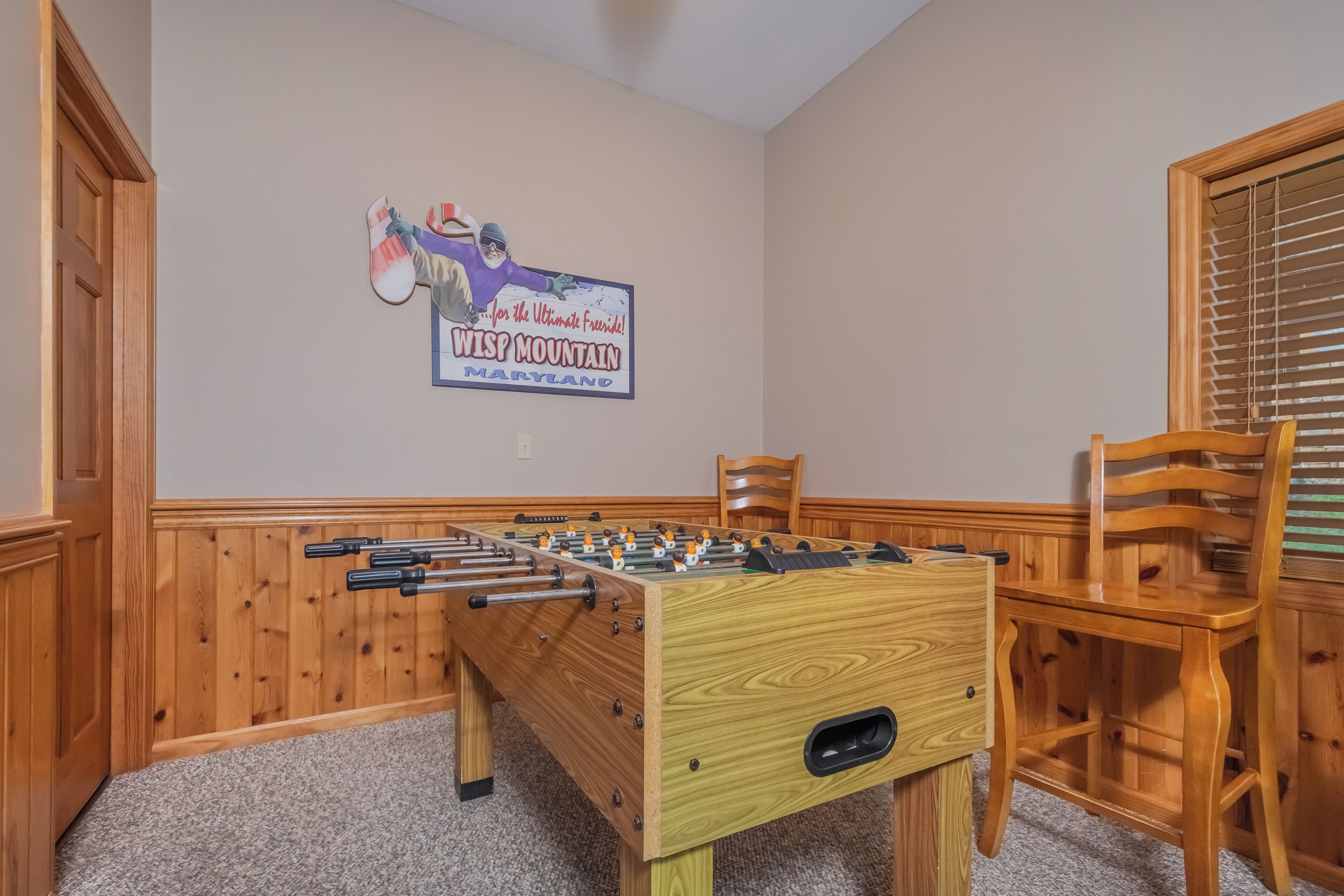 A wooden foosball table with a sign above it, wooden chairs, and a window with blinds.