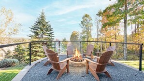 Un foyer entouré de quatre chaises Adirondack, installé sur une surface de gravier, avec une rambarde en métal et des arbres en arrière-plan.
