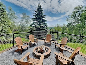 A deck with wooden chairs arranged around a fire pit, surrounded by trees and a view of a mountain range. - Green Gables by Taylor Made Deep Creek (McHenry)