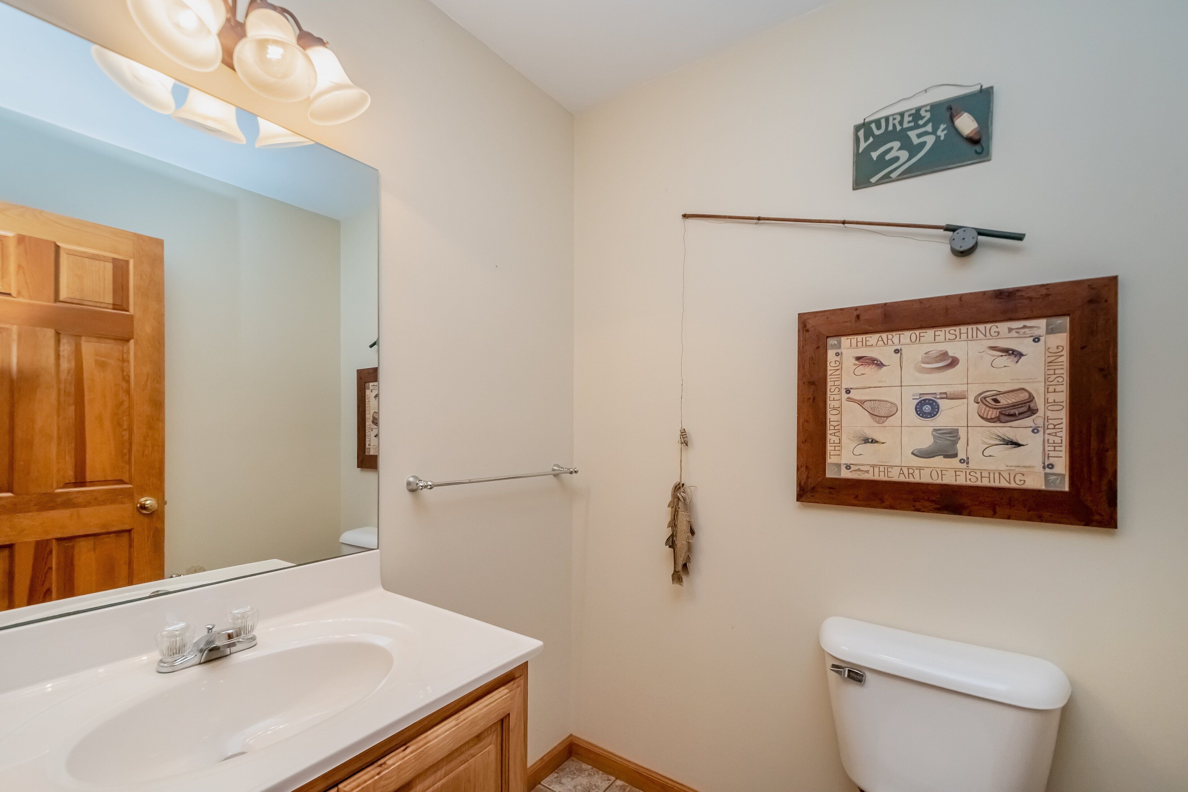 A bathroom with a wooden door, a white sink, a mirror, a towel rack, a toilet, and a fishing-themed wall decoration.