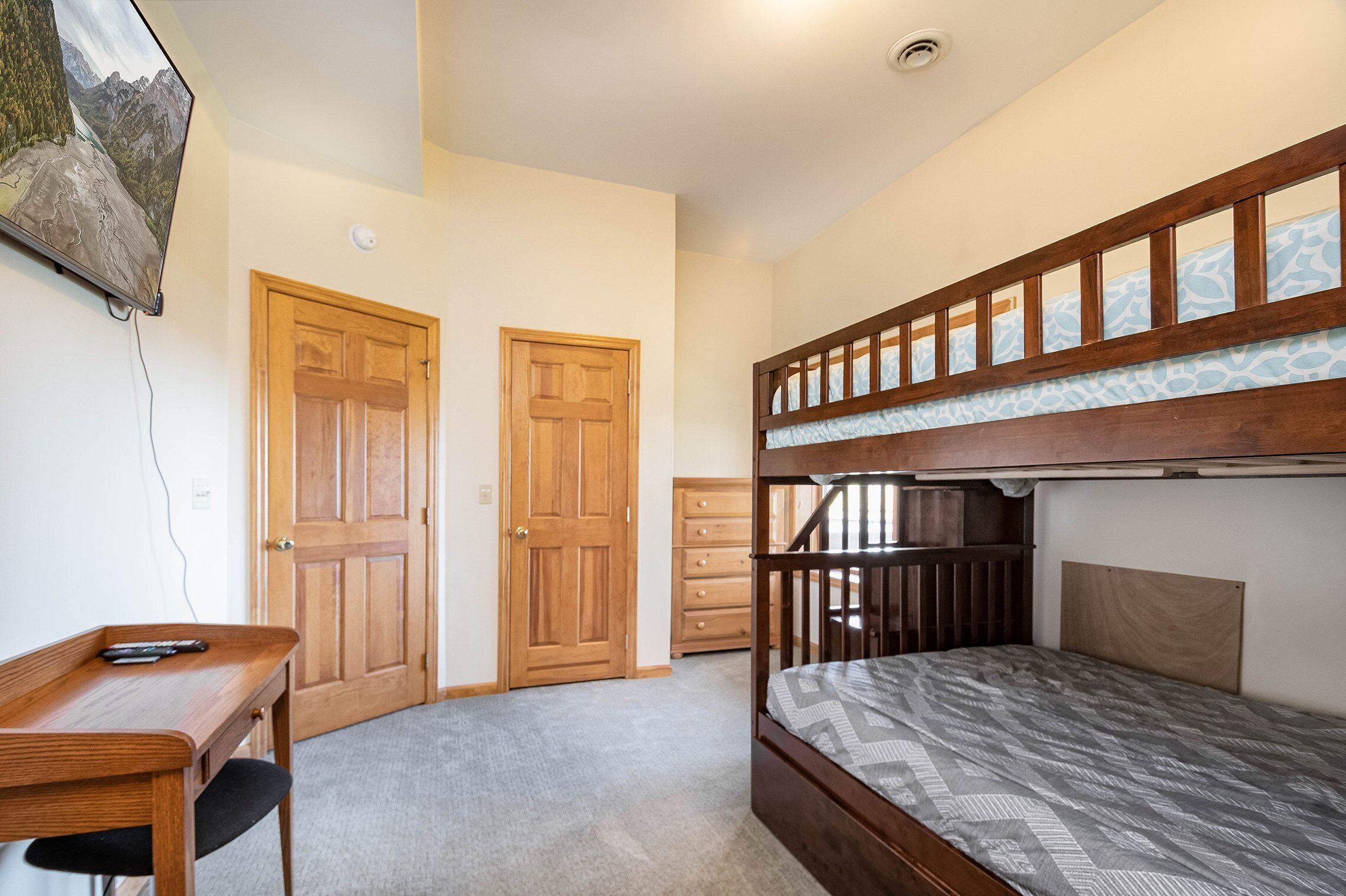 A bedroom with a bunk bed, a desk with a chair, two wooden doors, and a dresser.