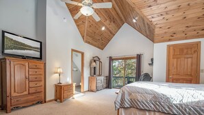Une chambre avec un grand lit, un plafond en bois, un téléviseur posé sur une commode et une vue sur l’extérieur par une porte.