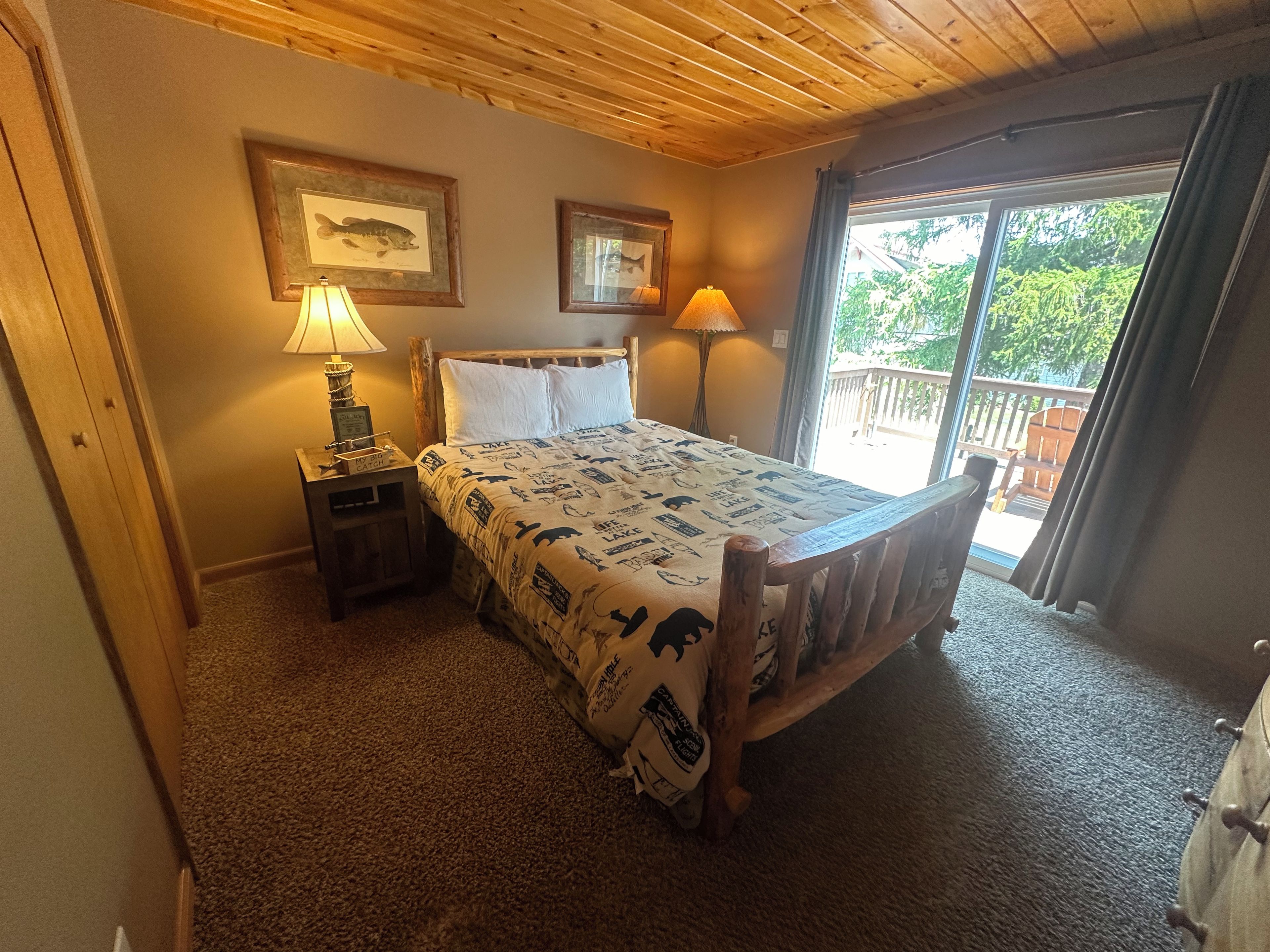 A bedroom with a wooden ceiling, a bed with a patterned bedspread, two framed pictures on the wall, a nightstand with a lamp, and a window with a view of greenery.
