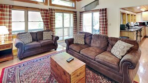 A spacious living room with two brown leather sofas, a wooden coffee table, and a large window with plaid curtains. - Happy Trails by Taylor Made Deep Creek (Oakland)