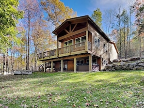 A two-story log cabin with a balcony, surrounded by trees and a stone wall. - Happy Trails by Taylor Made Deep Creek (Oakland)