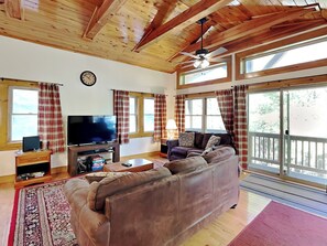 A living room with a large brown sofa, a television, and a wooden ceiling. There are windows with curtains and a clock on the wall.