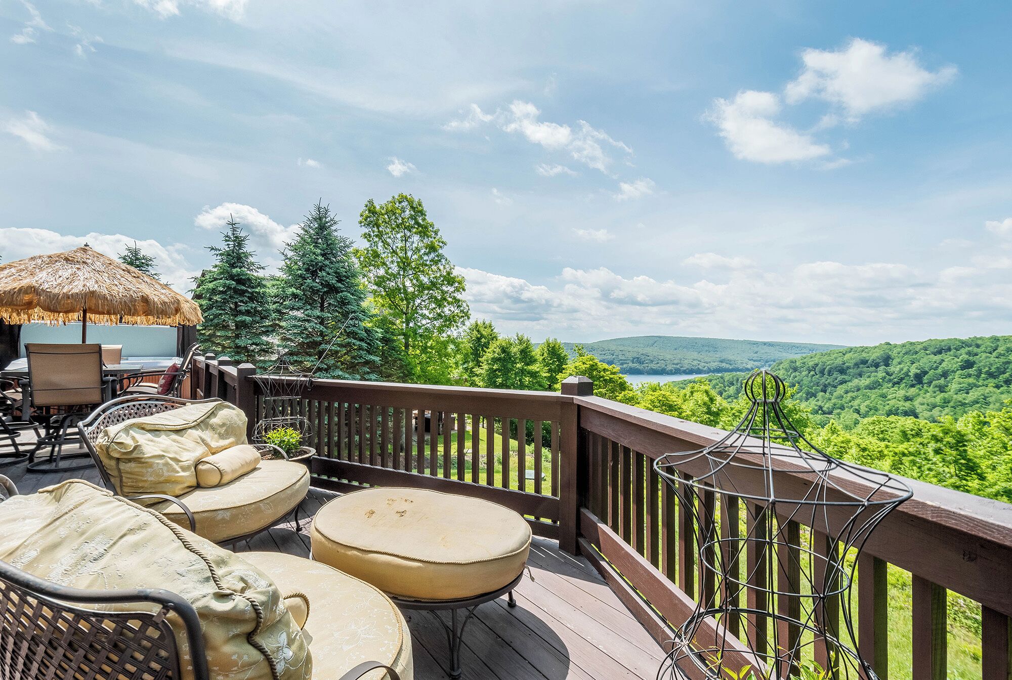 Una terraza con mobiliario exterior, un parasol de paja y una vista panorámica de un paisaje boscoso.