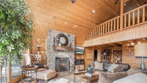 A cozy cabin interior with a stone fireplace, wooden ceiling, and a balcony.