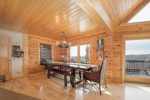 A wooden cabin interior with a dining table, benches, a chandelier, and large windows offering a view of the outdoors. - Happyplace Hideaway by Taylor Made Deep Creek (McHenry)