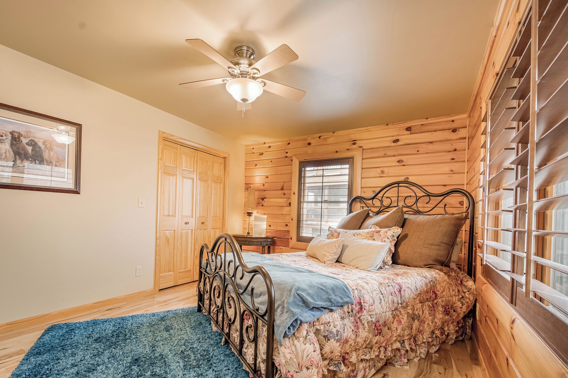 Une chambre avec un plafond en bois, un lit recouvert d’une literie à motifs floraux, un tapis bleu, un ventilateur de plafond et une fenêtre avec des stores.