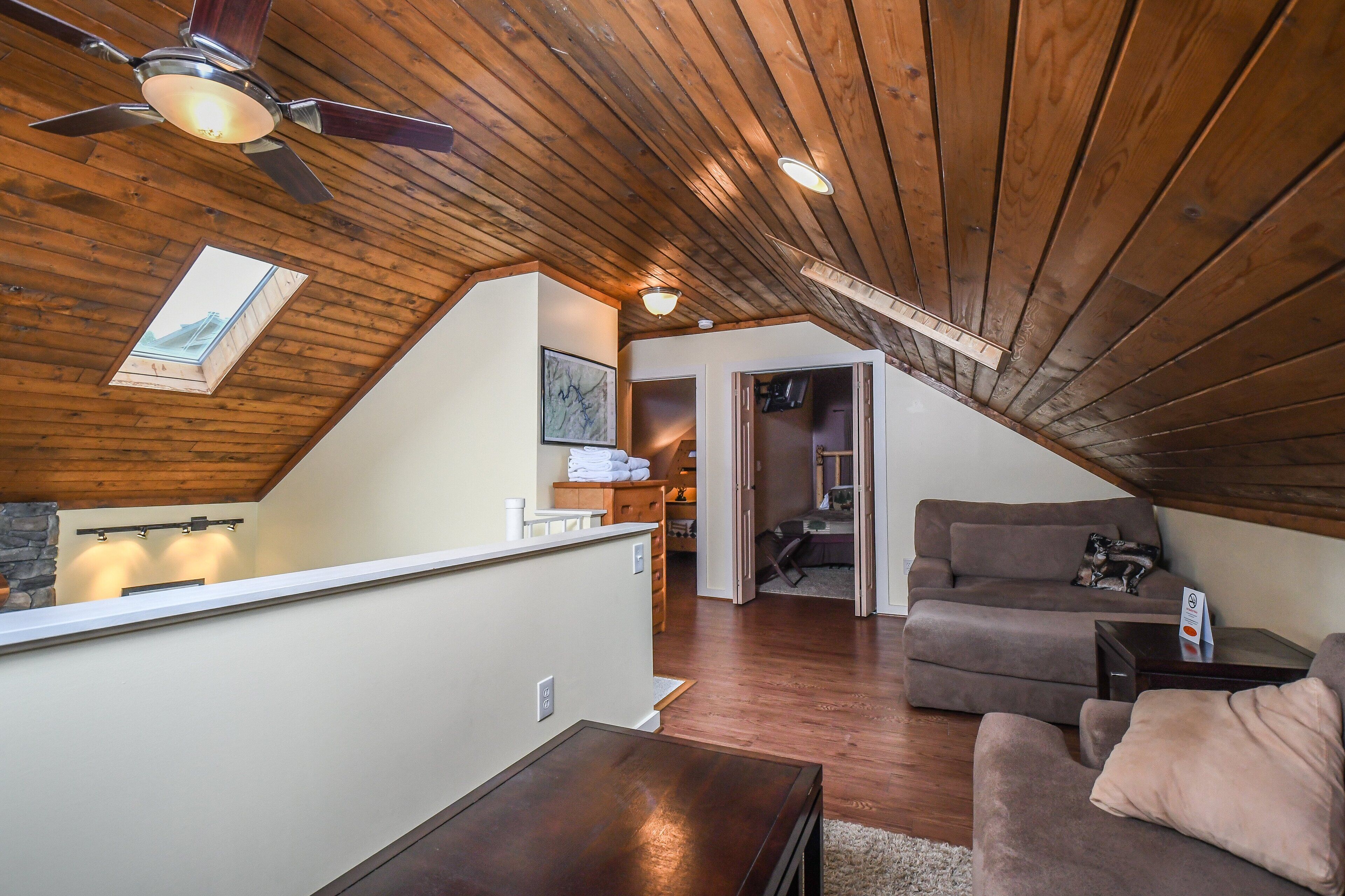 A loft-style living space with a sloped ceiling, wooden beams, a ceiling fan, a staircase, a couch, a coffee table, and a small kitchen area.