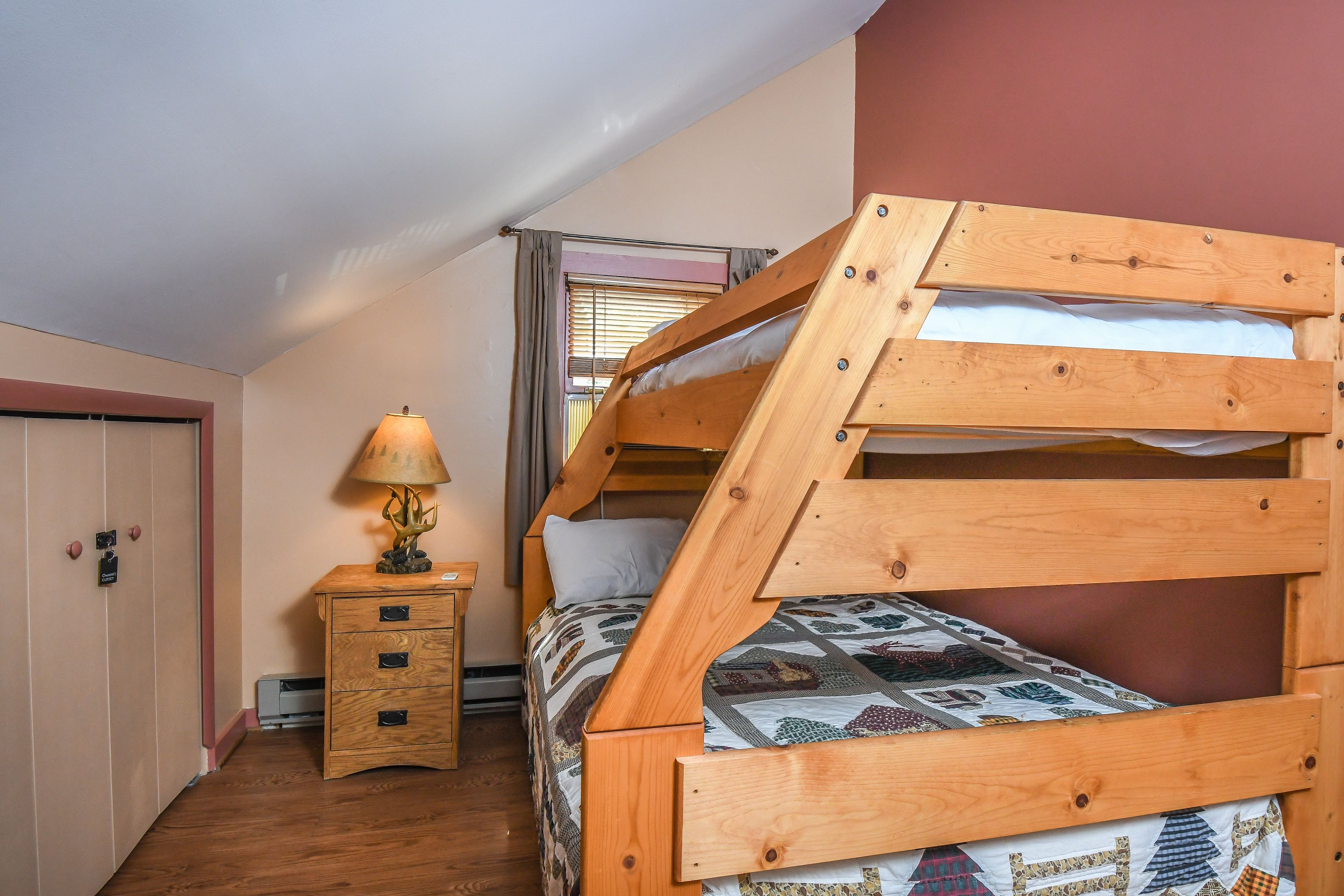 A bedroom with a wooden bunk bed, a bedside table with a lamp, and a window with blinds.