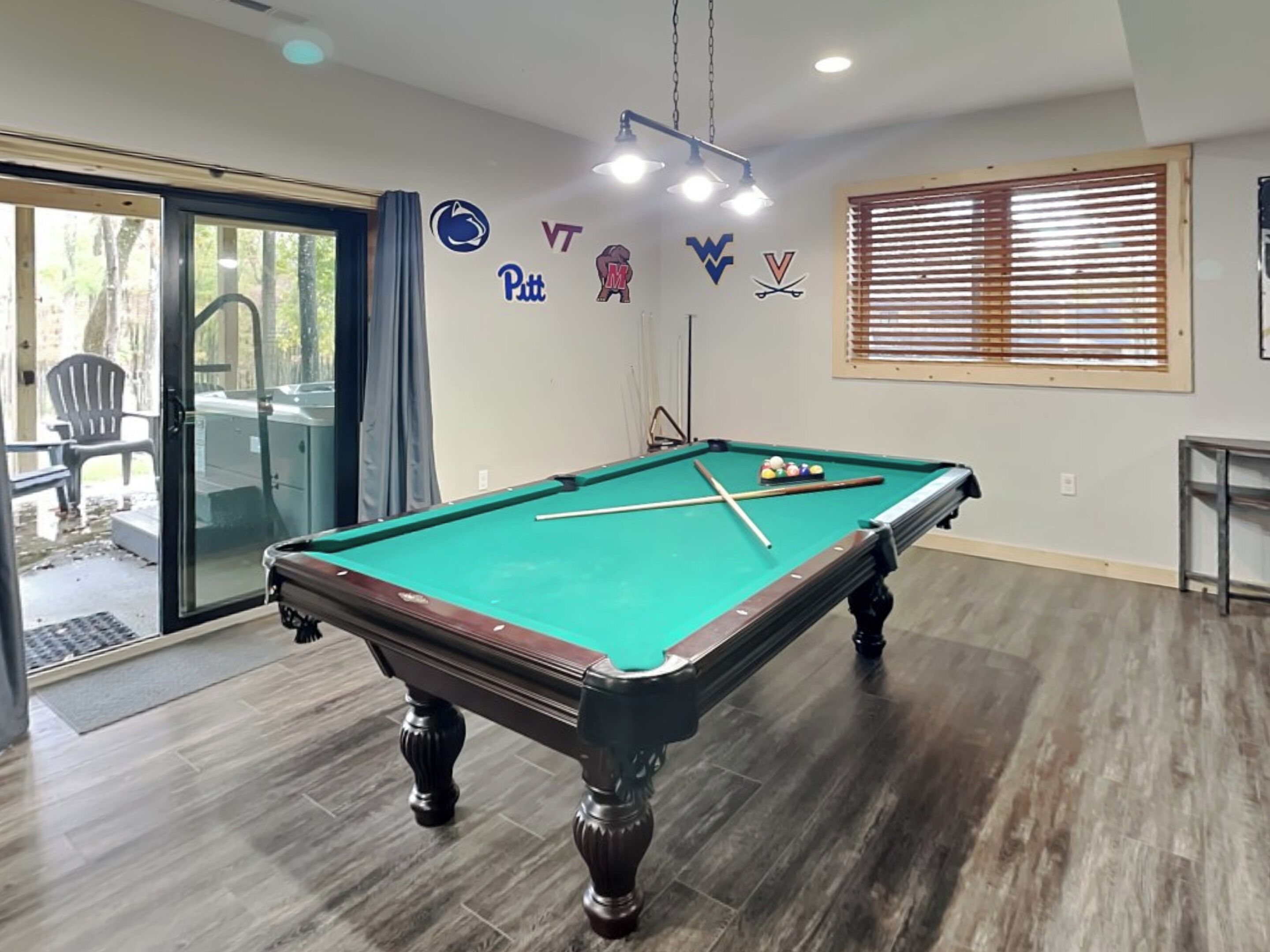 A pool table with a green felt surface, surrounded by wooden flooring. There are sports team logos on the wall, and a window with blinds to the right.