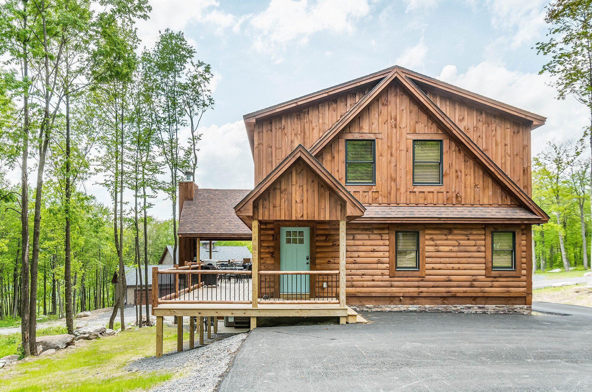 A two-story log cabin with a porch and a deck, surrounded by trees.