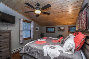 Ein Schlafzimmer mit Holzbalkendecke, einem Bett mit floralem Bettzeug, einem Deckenventilator, einem Schminktisch und einem an der Wand befestigten Fernseher.