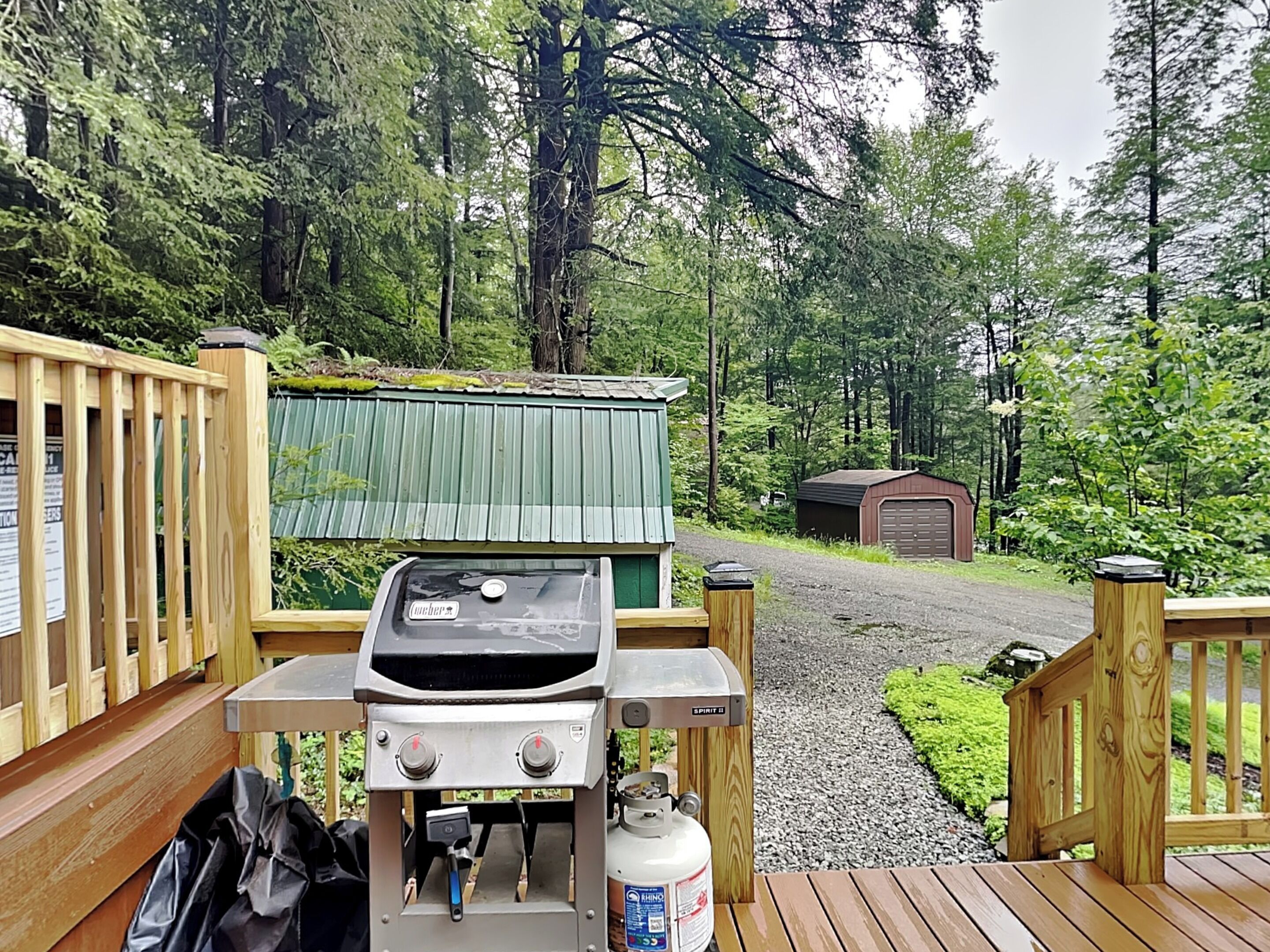 A wooden deck with a barbecue grill and propane tank, surrounded by a forested area with a small shed and a gravel path.