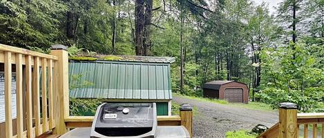 A wooden deck with a barbecue grill and propane tank, surrounded by a forested area with a small shed and a gravel path.