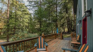 Eine Holzterrasse mit Adirondack-Stühlen und einem Tisch, umgeben von einem Wald.