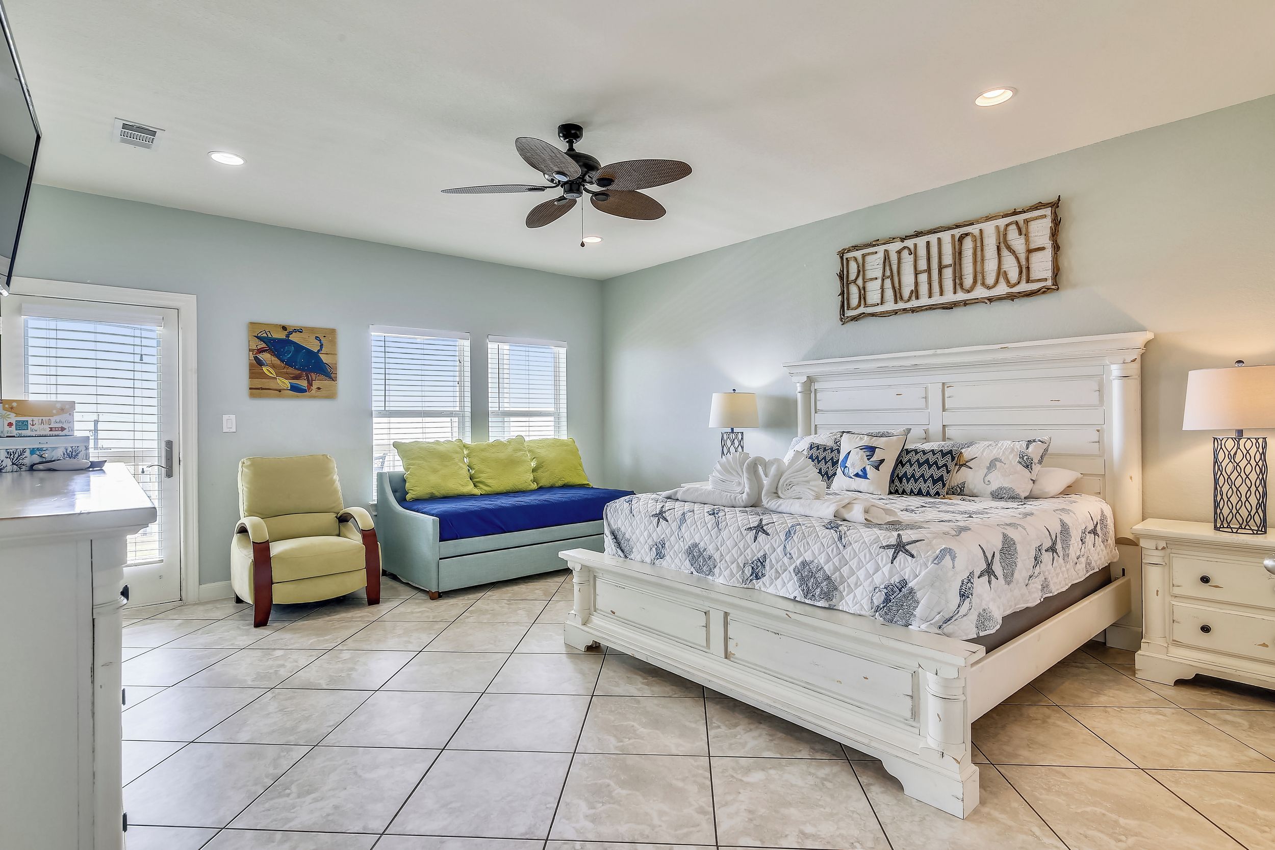 A bedroom with a large bed, a ceiling fan, a chair, a sofa, and a sign that reads "BEACHHOUSE".
