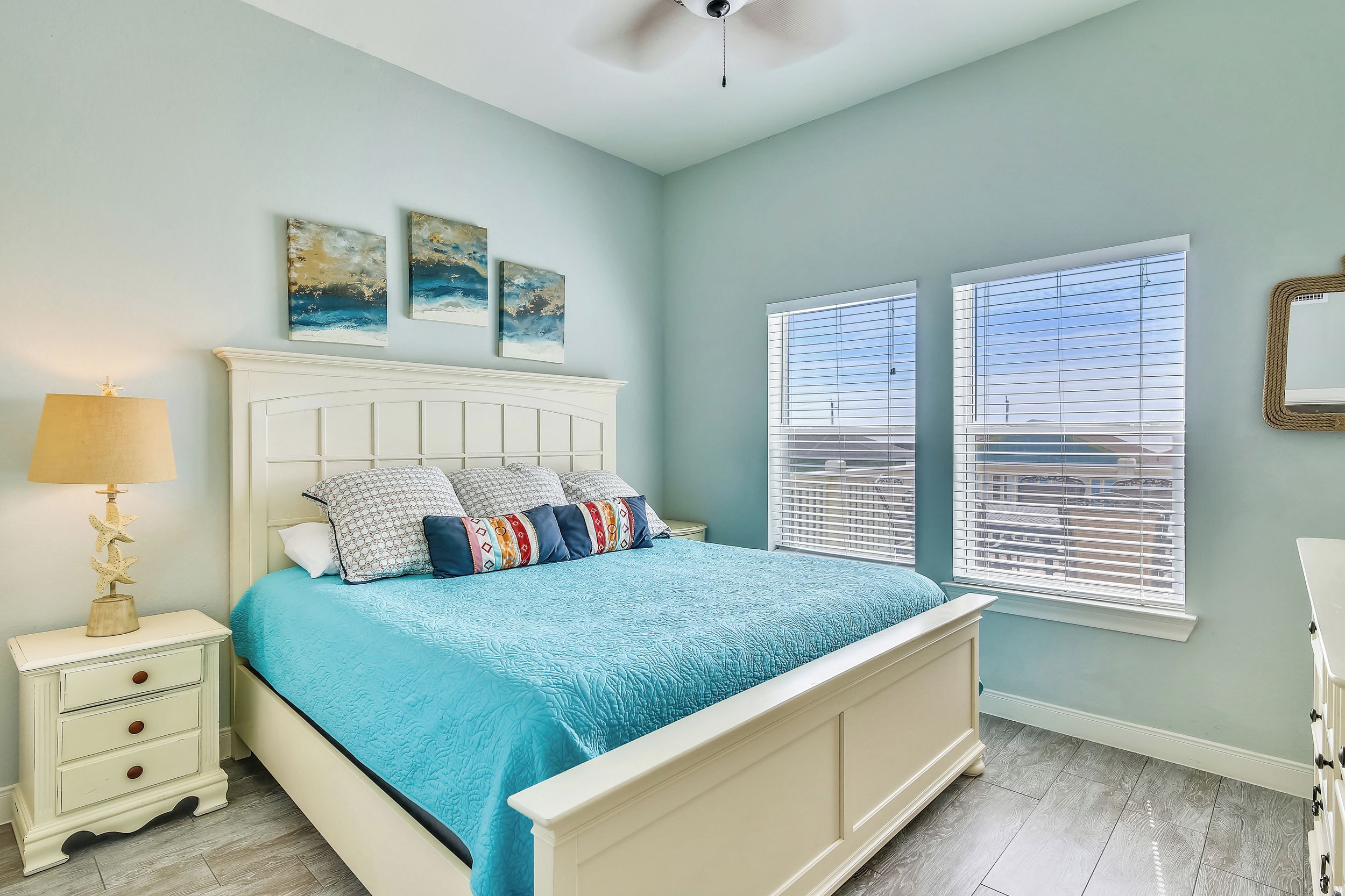 A bedroom with a bed, nightstand, lamp, artwork, and windows.