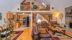 A spacious living area with a high ceiling, wooden furniture, a fireplace, and a kitchen in the background. - Heart and Soul by Taylor Made Deep Creek (Oakland)