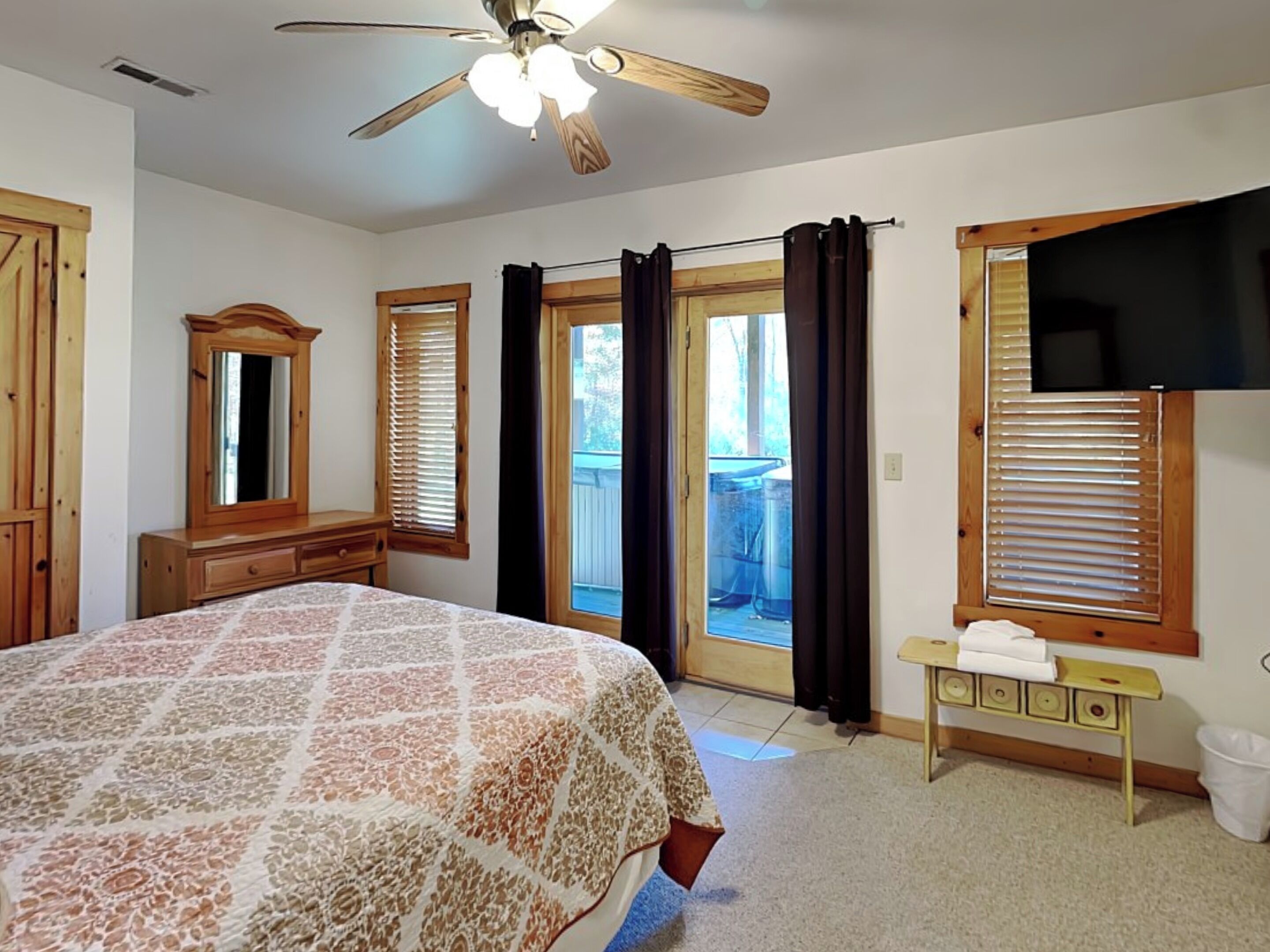 Une chambre à coucher avec un grand lit, un ventilateur de plafond, une commode en bois, un miroir et une fenêtre avec des stores.