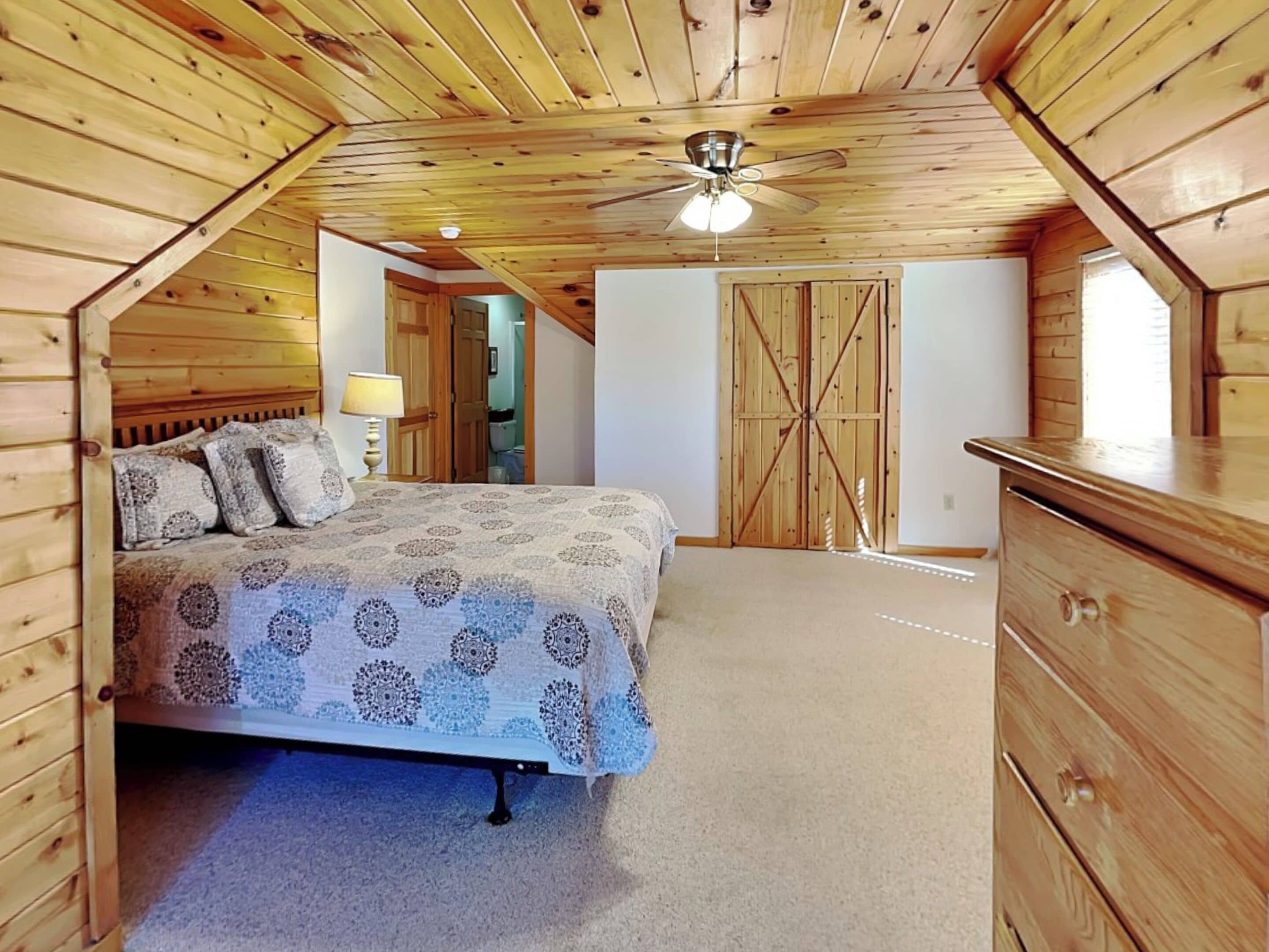 Un dormitorio con techo de madera, una cama con ropa de cama estampada, un aparador y una puerta que da a otra habitación.