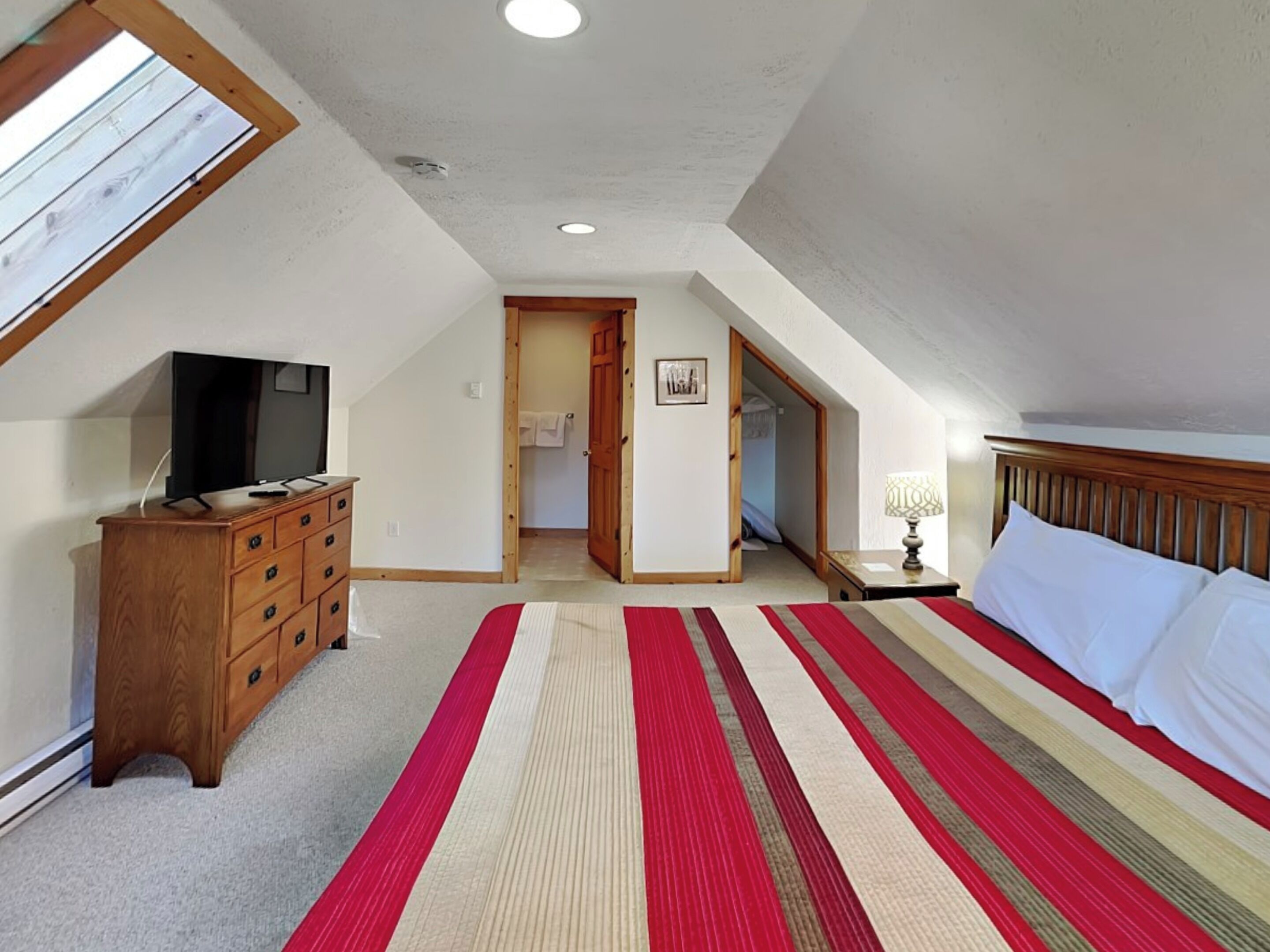 A bedroom with a sloped ceiling, a wooden dresser, a television, a bed with a striped rug, and a hallway leading to a bathroom.